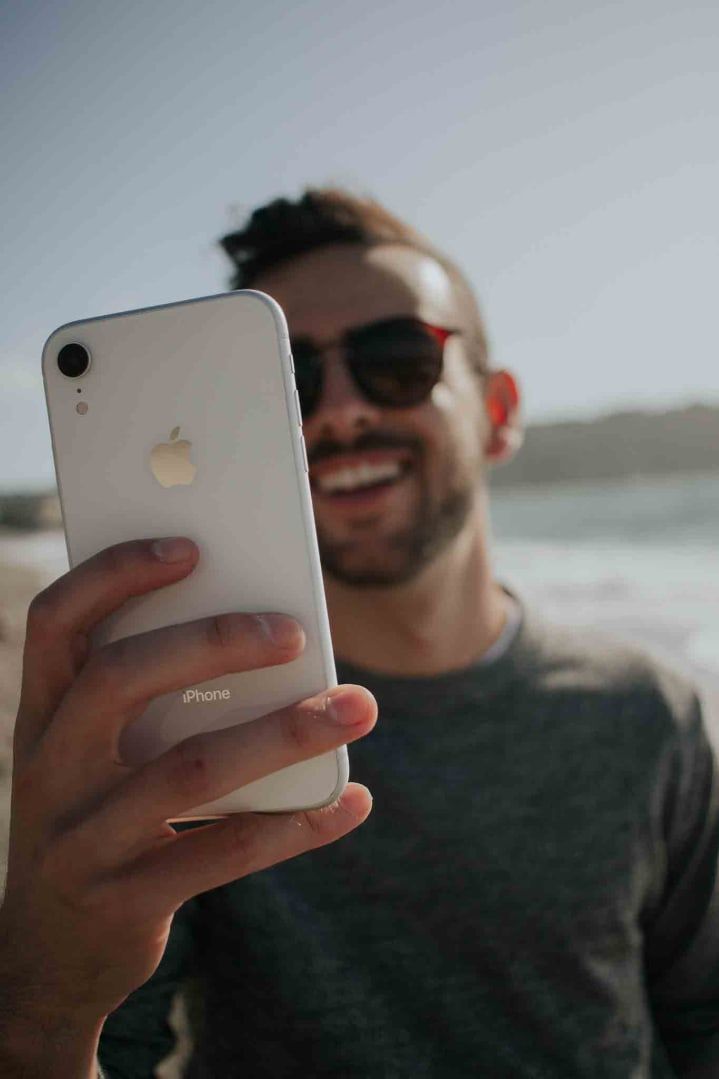 Smiling man in sunglasses holding up an iPhone by the water