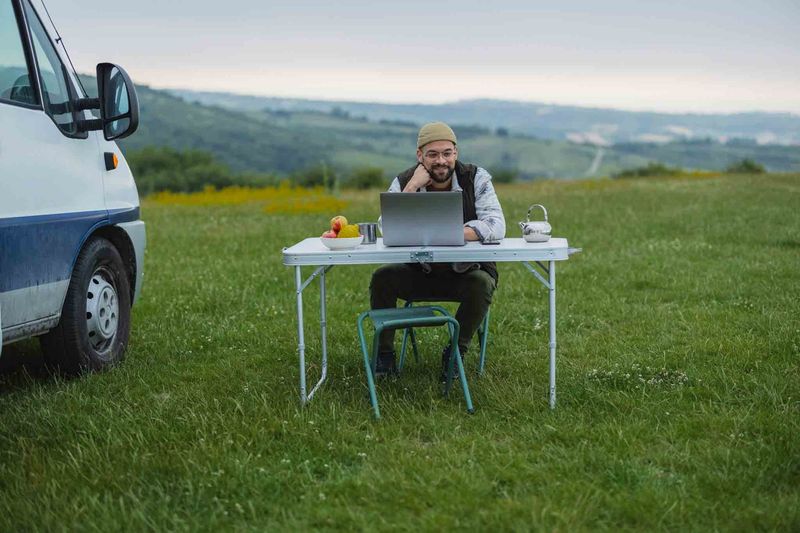 Rental owner on a laptop at a table in a field next to a van – supporting self-service customers remotely