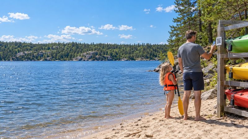 Father scanning the QR code on a self-service kayak rack at a lake beach to book on his phone, his daughter in a life jacket beside him