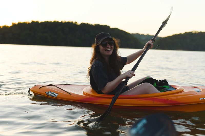 Smiling woman in a cap and sunglasses paddling an orange kayak on a lake