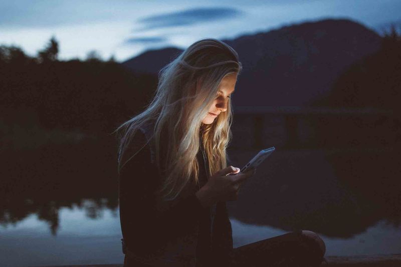 Woman using her phone by a lake at dusk, mountains in the background